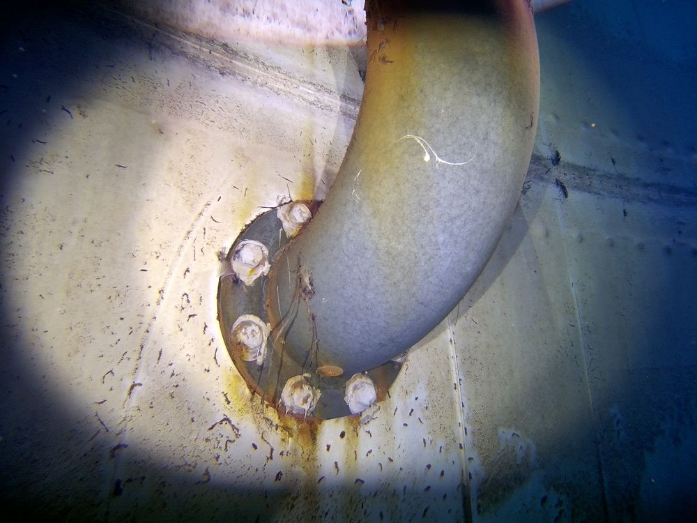 A pitted, rust-stained pipe elbow and flange on the floor of a steel water tank, seen by the ROV.