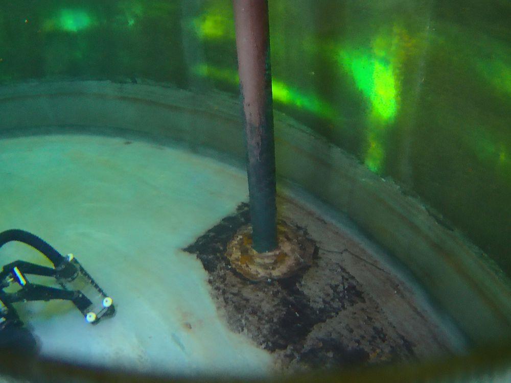 A robotic crawler at the base of a vertical pipe inside a reservoir, with a ring of sediment around the pipe.