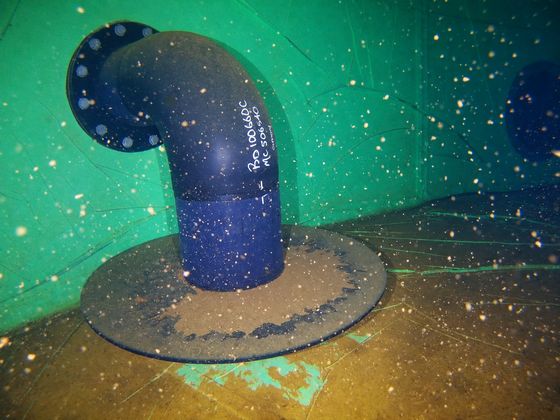 A blue pipe elbow rising from a sediment-covered tank floor, photographed underwater by the ROV.