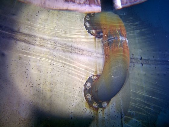 A pipe elbow entering through a riveted steel tank wall, photographed underwater with the ROV lights.