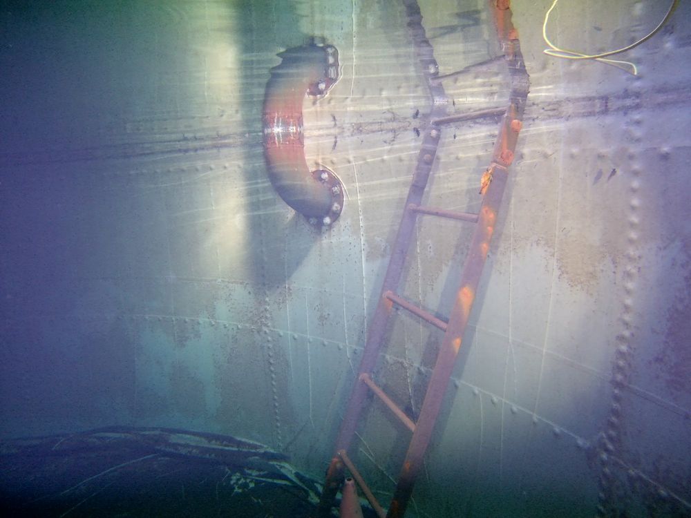 An internal ladder and inlet elbow inside a steel water tank, with corrosion staining on the wall panels.