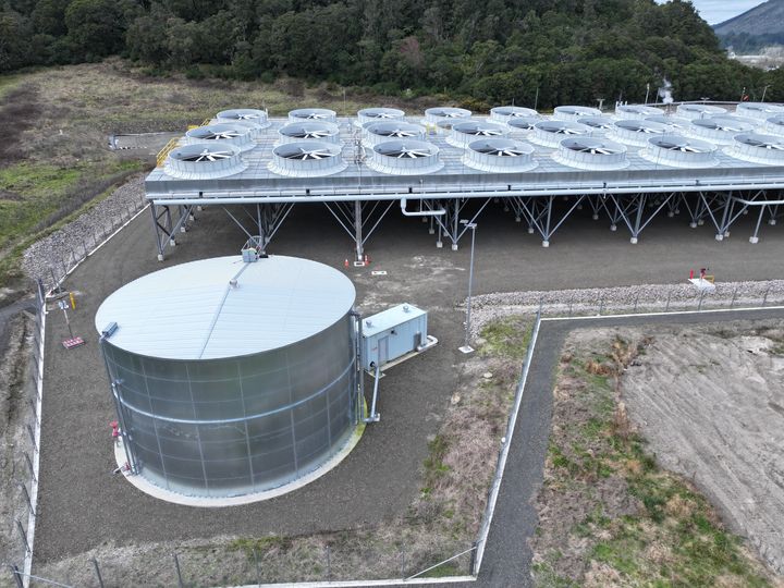 A galvanised steel water tank in a fenced compound beside a bank of industrial cooling fans.