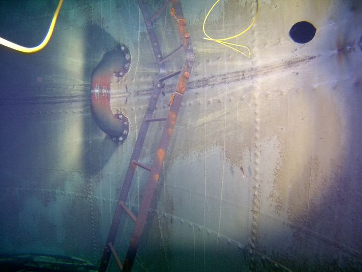 A rusted internal ladder and a pipe elbow inside a steel water tank, photographed underwater with the ROV tether visible.